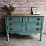 An image showcasing a skilled hand lightly applying Americana Decor Chalk Paint onto a vintage wooden dresser, revealing its vibrant, matte finish